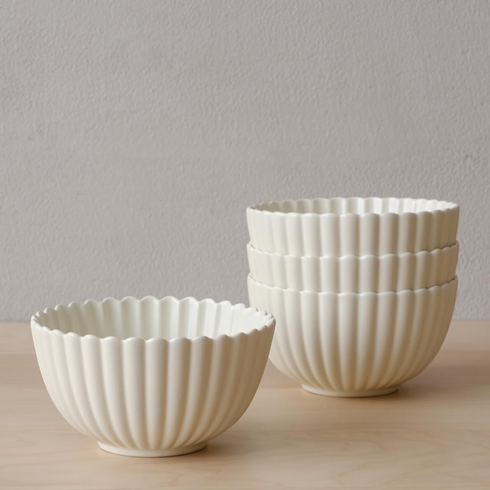 Four white ceramic bowls with fluted, scalloped edges are arranged on a light wooden surface against a plain gray wall—perfect for serving your favorite dinner party recipes. Three bowls are stacked, and one stands alone.