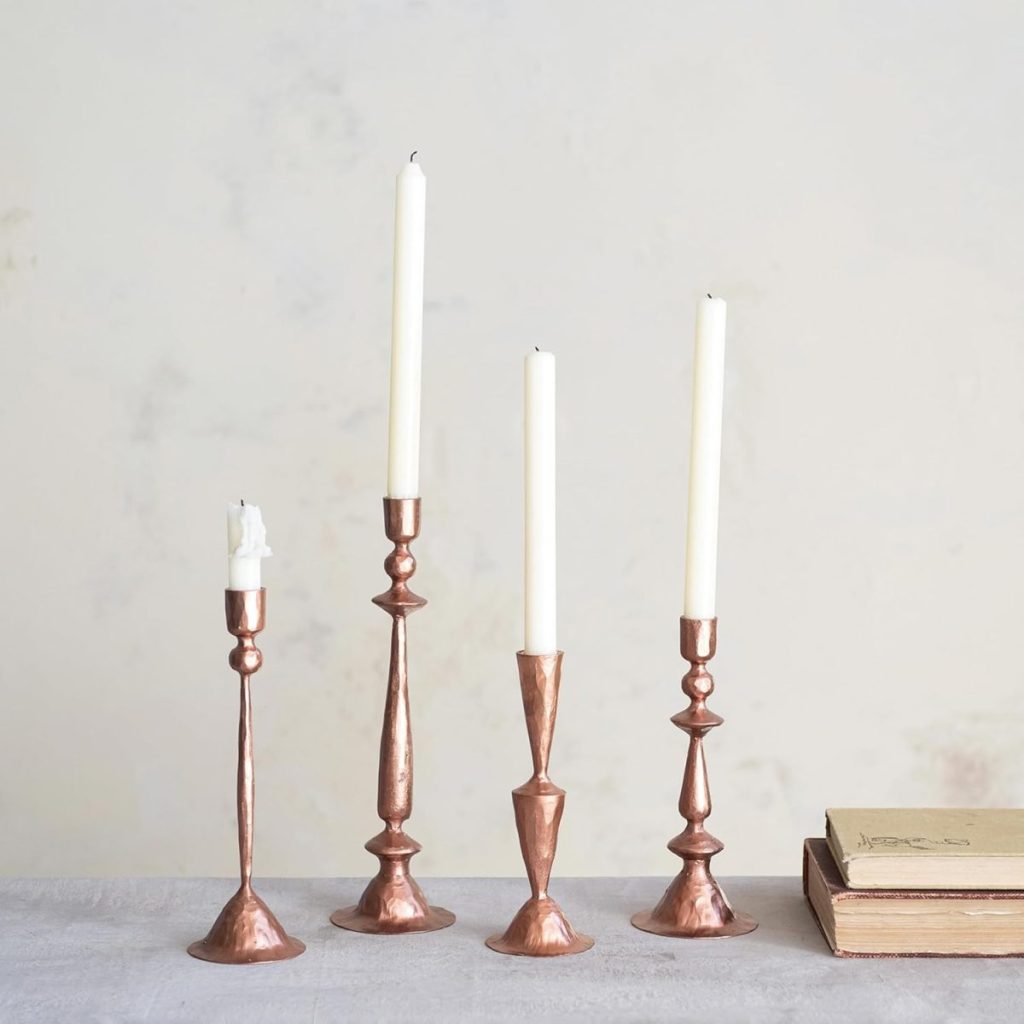 Four tall copper candlesticks with white candles, one partially melted, are arranged on a gray surface next to two closed books against a light background—perfect inspiration for elegant outdoor table decor.