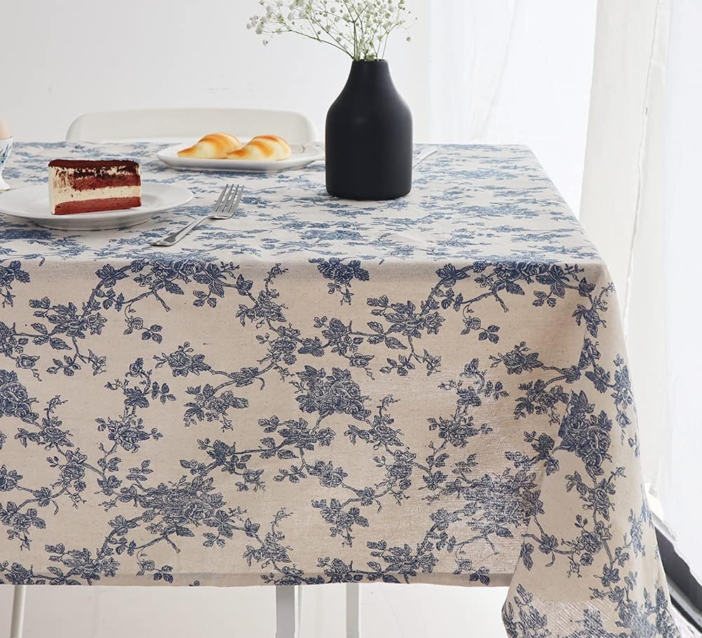 A charming example of outdoor table decor, this setup features a blue floral-patterned tablecloth, a black vase with white flowers, a plate of cake, a fork, and tempting pastries.