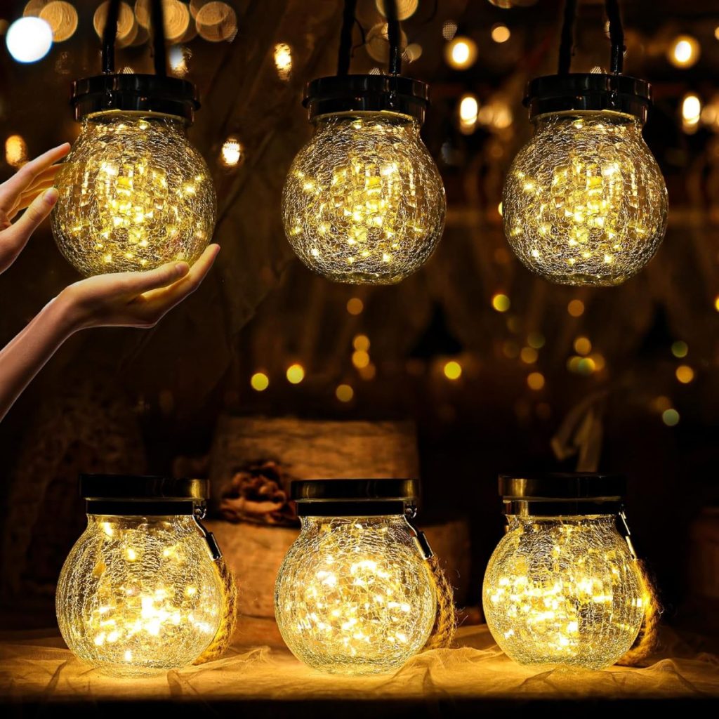 Six glass jar lanterns filled with warm white fairy lights are displayed as enchanting outdoor table decor, with a hand reaching toward one of the hanging lanterns.