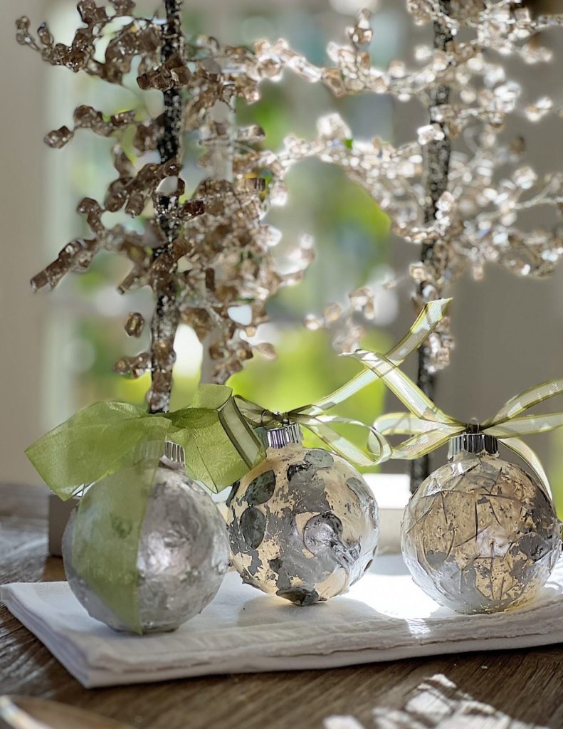 Three metallic Christmas ornaments with green and yellow ribbons rest on a folded white napkin, making for festive Christmas in July ideas, with a decorative branch in the background.