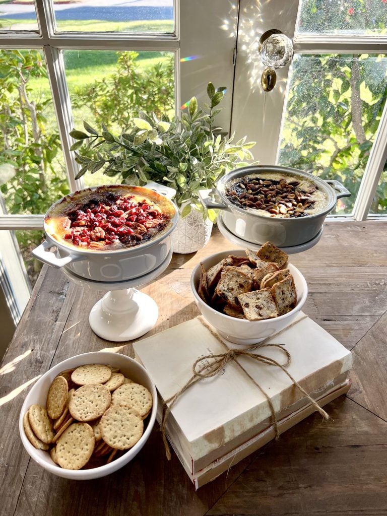 Two baked brie appetizers on small cake stands with two small bowls of crackers, one on top of a stack of books tied with twine. Small green plant in background with french doors behind