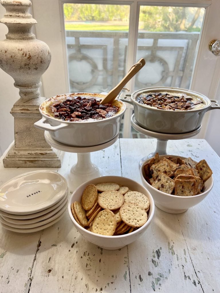 Two baked brie appetizers on small cake stands with two small bowls of crackers and small Rae Dunn nibble plates. Farmhouse lamp in background