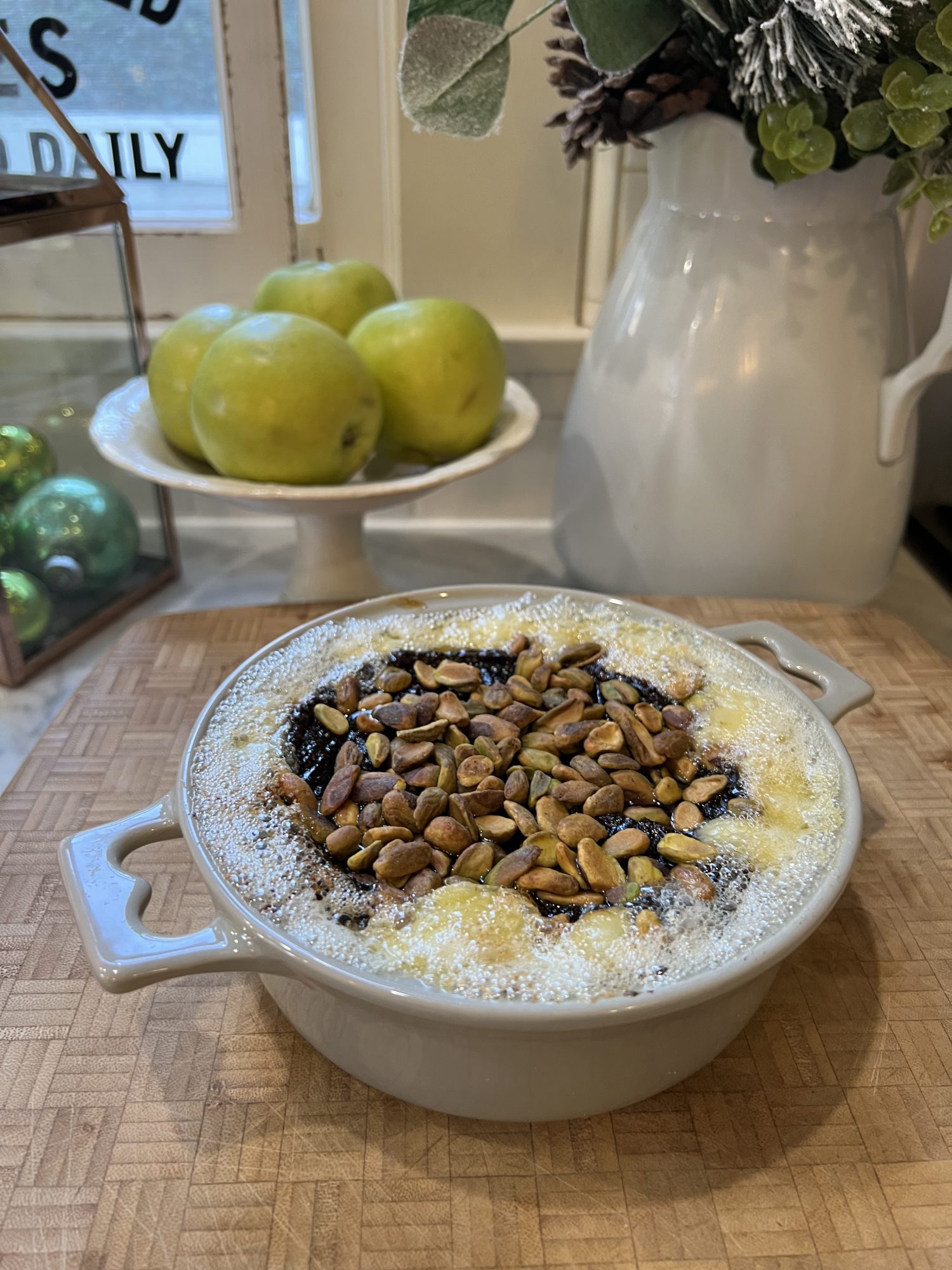 Baked brie with fig jam and pistashios right out of oven, bubbly and warm. Background, white pitcher with greenery, green apples on footed plate