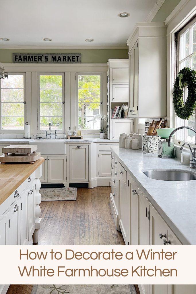 a white kitchen with windows and an island with a bowl with apples, a cutting board and wreaths in the windows.