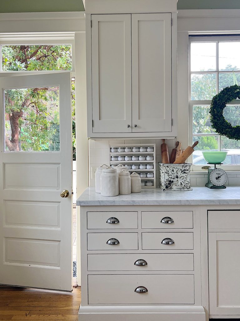 a white kitchen with windows and an island with a bowl with apples, a cutting board and wreaths in the windows.