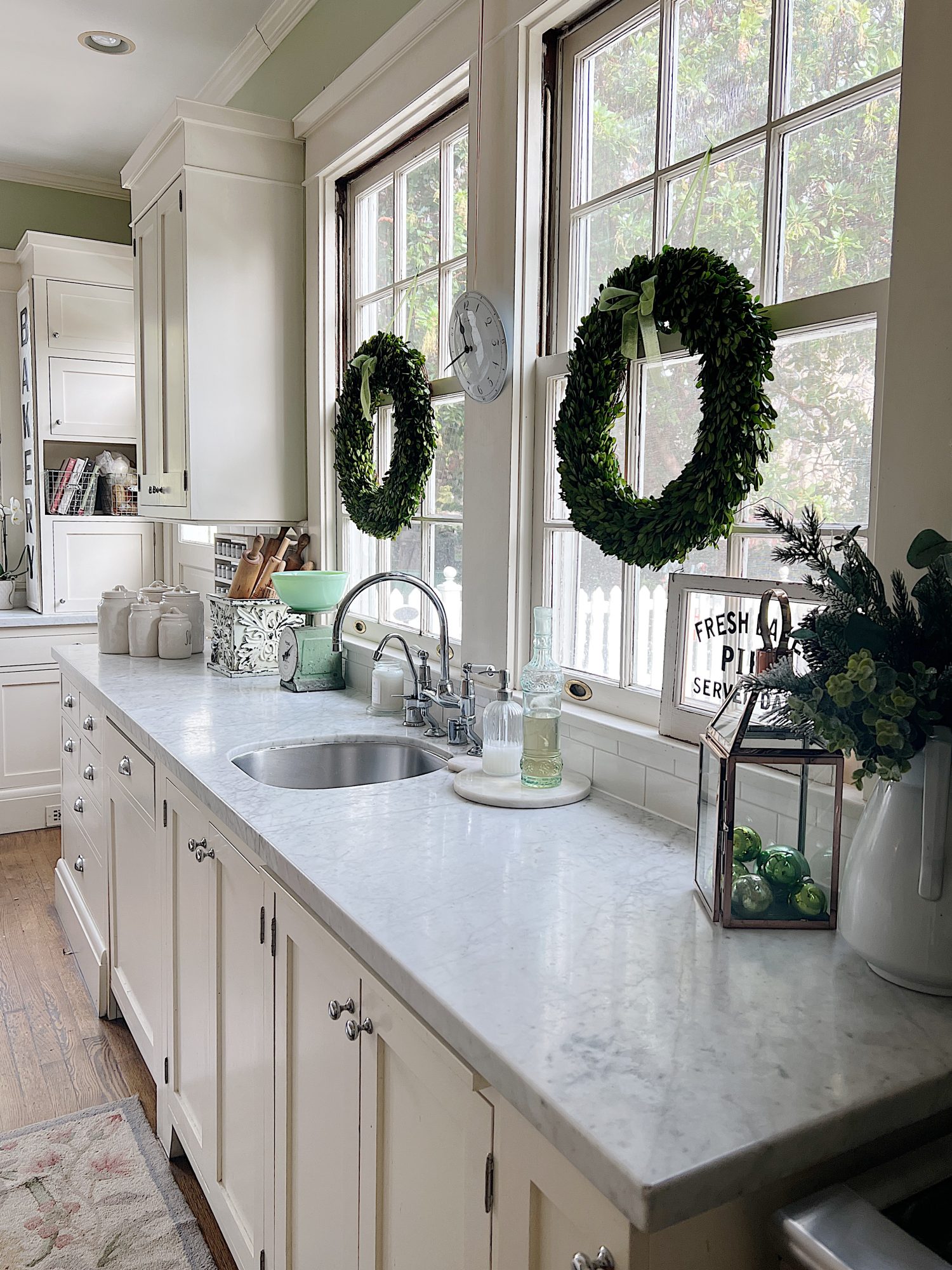 a white kitchen with windows and an island with a bowl with apples, a cutting board and wreaths in the windows.