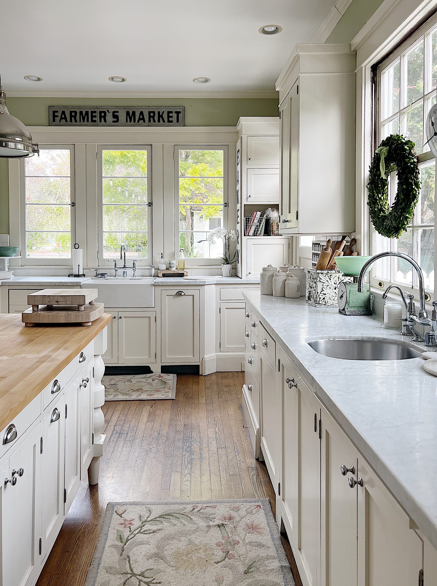 a white kitchen with windows and an island with a bowl with apples, a cutting board and wreaths in the windows.