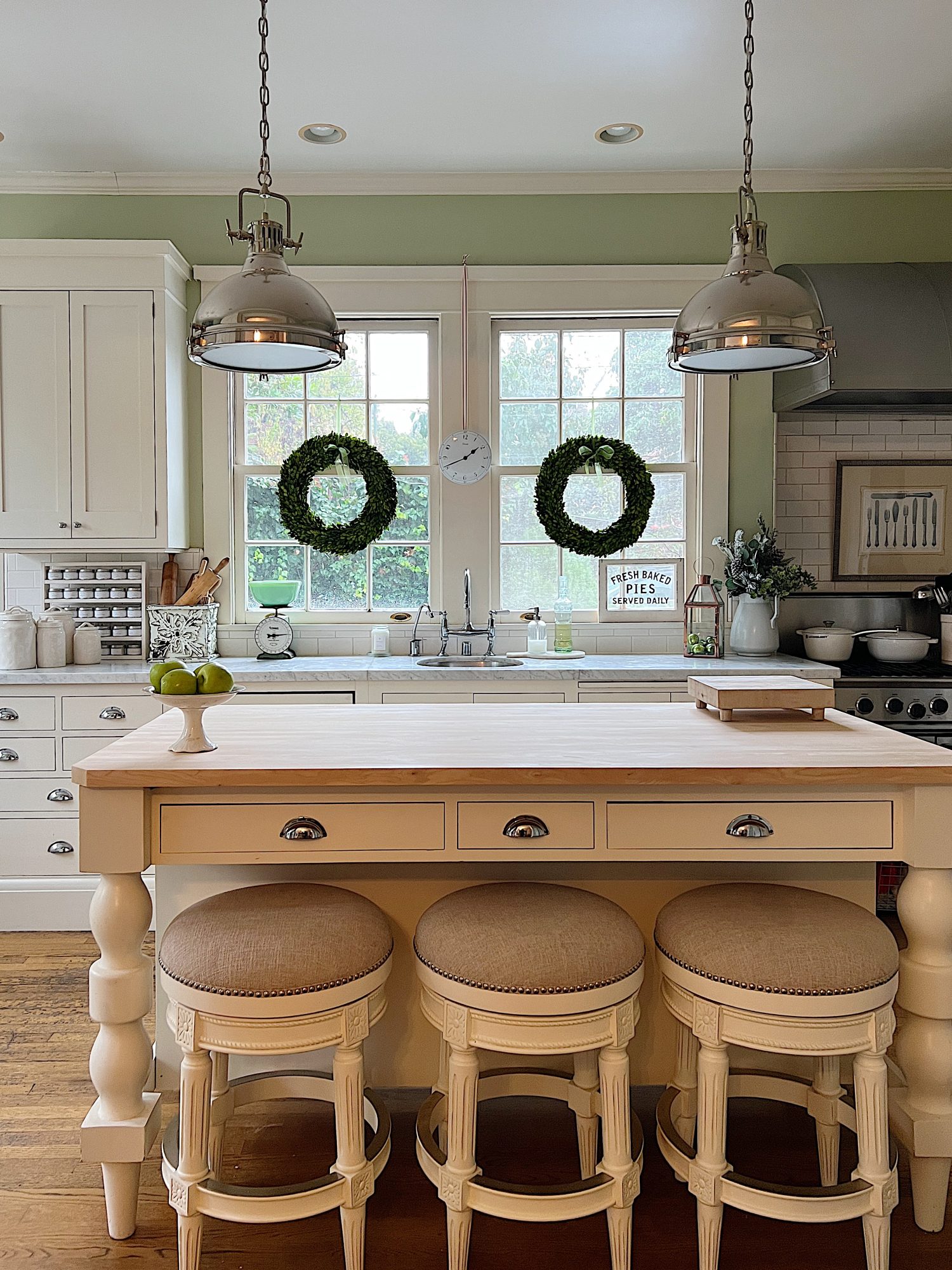 a white kitcehn with windows and an island with a bowl with apples, a cutting board and wreaths in the windows.