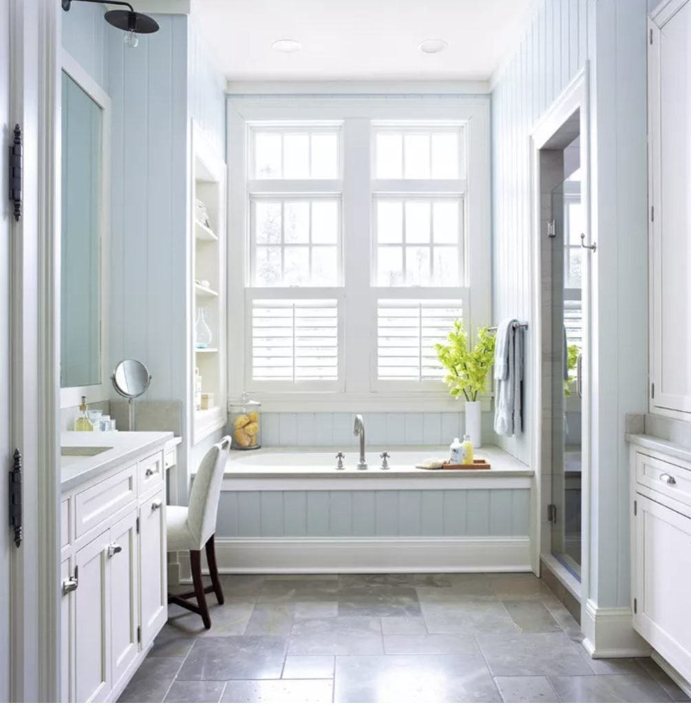 Bathroom with sky blue painted ship lap. Bathtub underneath tall french paned windows with shutters and shelves to one side filled with decor. Sink and vanity are in view to the left side of the tub, shower and linen closet on right hand side.