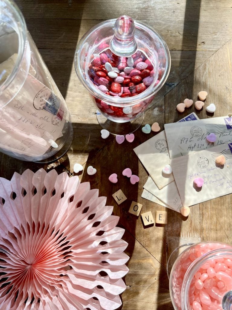 Top down view of table with apothecary jars filled with vintage letters and candy. Scatterd on table are candy conversation hearts, vintage letters, scrabble tiles LOVE and pink tissue fan