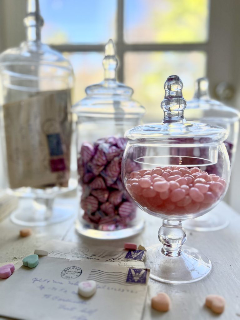 View of apothecary jars, one with vintage letters, three with candy. French window pane in background and vintage letters with scattered candy hearts on table