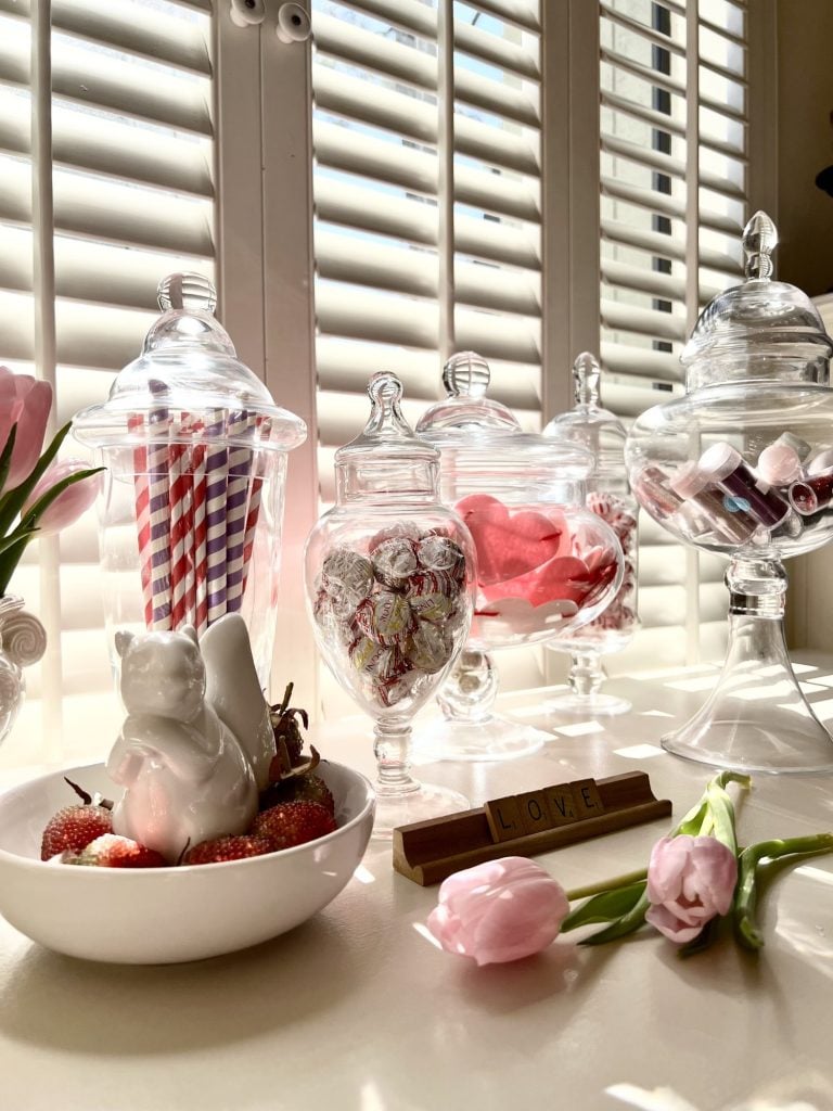 Glass apothecary jars filled with candies, red and purple striped paper straws, pink, red and white felt hearts and valentine colored tubes of glitter. Pink tulips on the table in front to scrabble tiles spelling LOVE