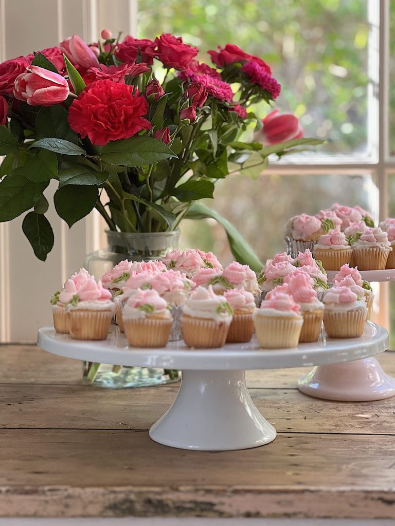 cupcakes decorated with white and pink buttercream frosting on two trays with a vase of flowers