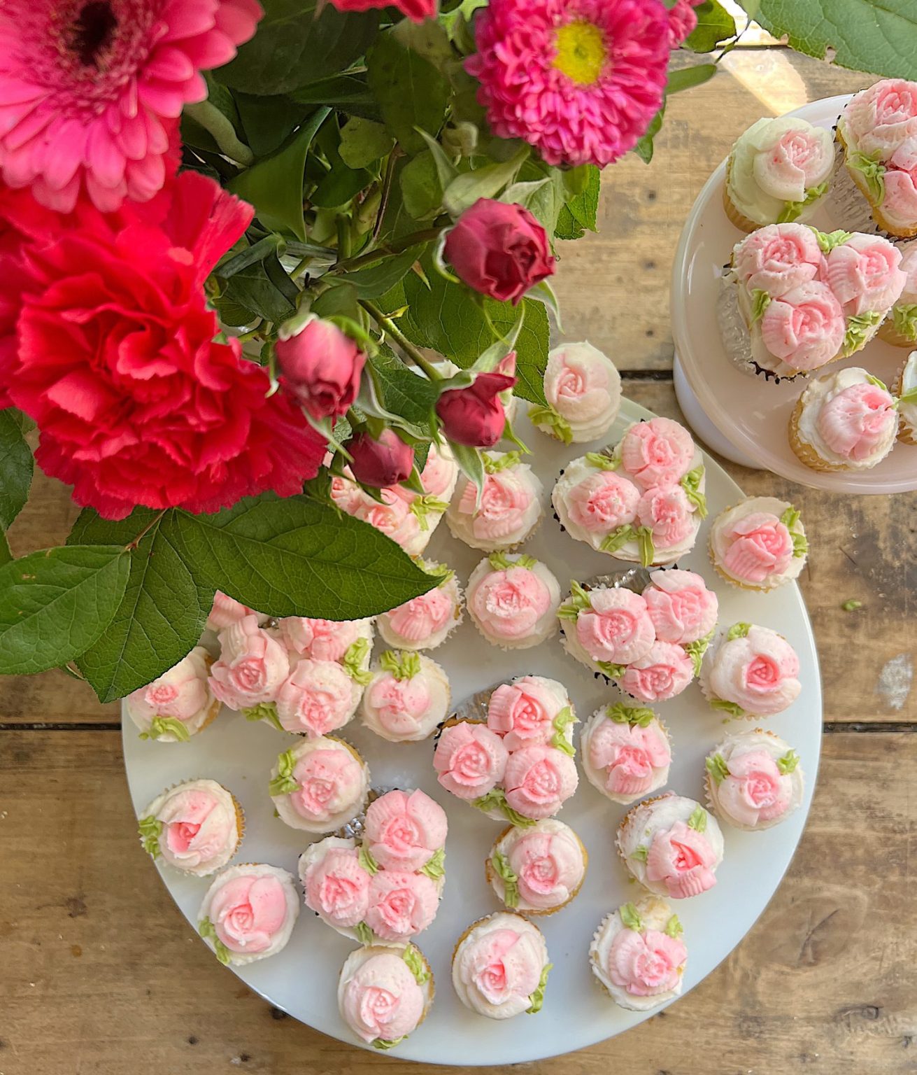 cupcakes decorated with white and pink buttercream frosting on two trays with a vase of flowers