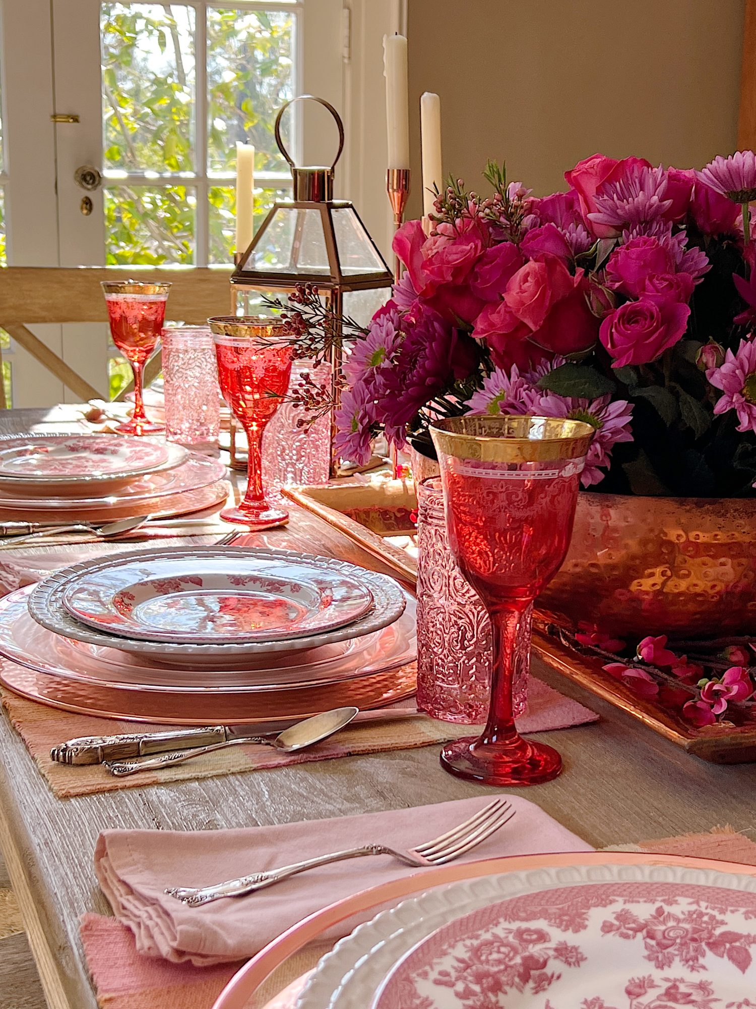 Valentine dining table with copper chargers, copper bowl, copper lantern, copper tray, and copper candlesticks. Pink water glasses and red wine glasses and fresh flowers are also on the table.