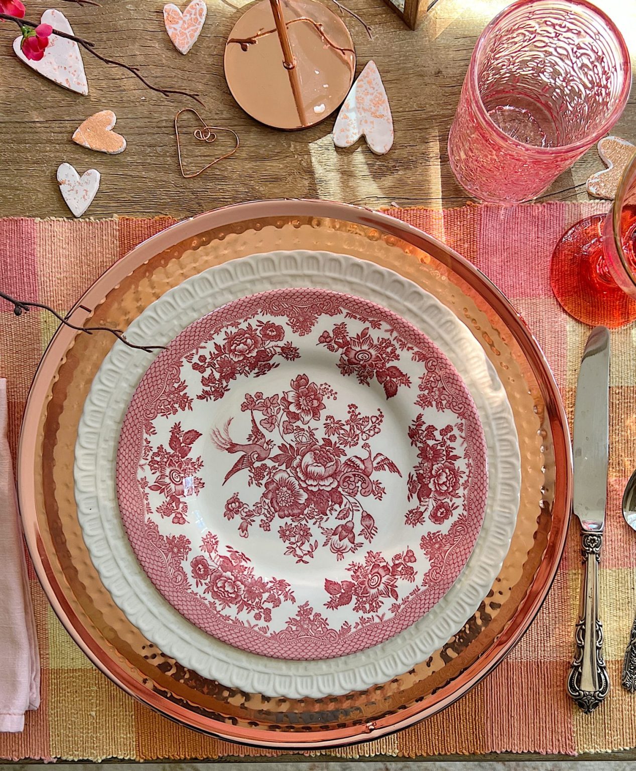 Valentine dining table with copper chargers, copper bowl, copper lantern, copper tray, and copper candlesticks. Pink water glasses and red wine glasses and fresh flowers are also on the table.