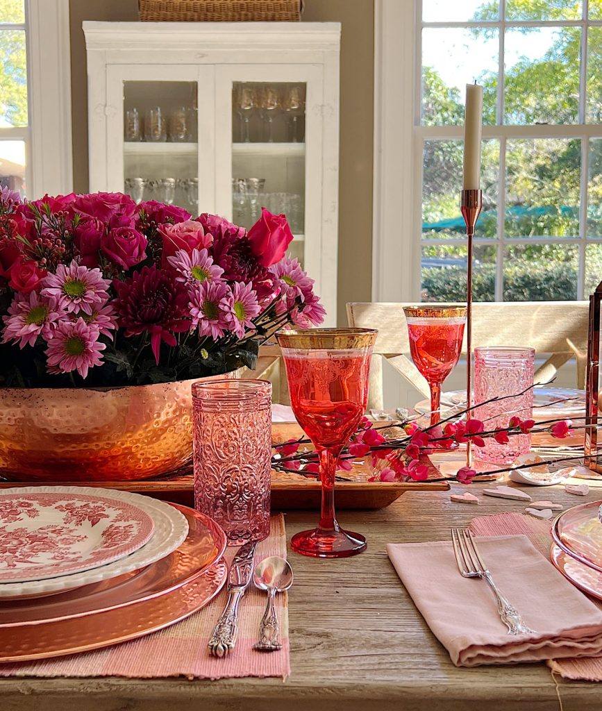 Valentine dining table with copper chargers, copper bowl, copper lantern, copper tray, and copper candlesticks. Pink water glasses and red wine glasses and fresh flowers are also on the table.