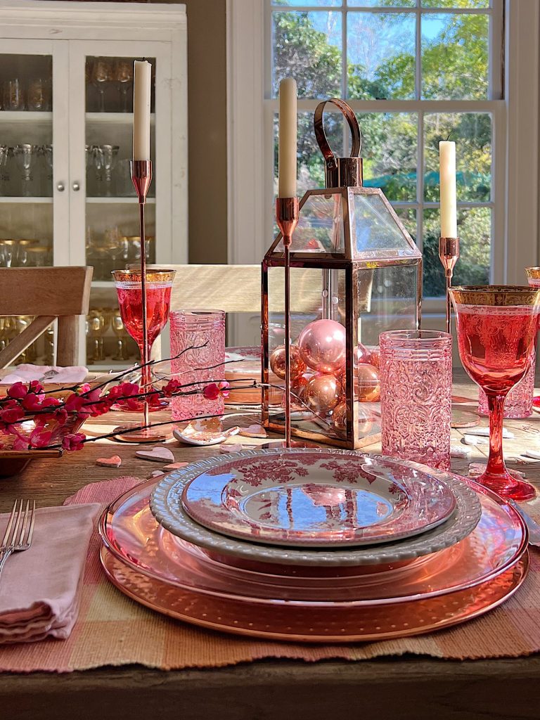 Valentine dining table with copper chargers, copper bowl, copper lantern, copper tray, and copper candlesticks. Pink water glasses and red wine glasses and fresh flowers are also on the table.