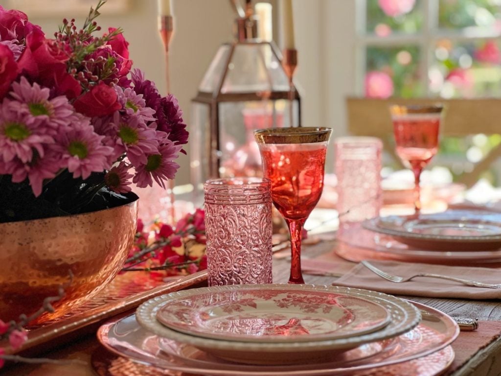 Valentine dining table with copper chargers, copper bowl, copper lantern, copper tray, and copper candlesticks. Pink water glasses and red wine glasses and fresh flowers are also on the table.