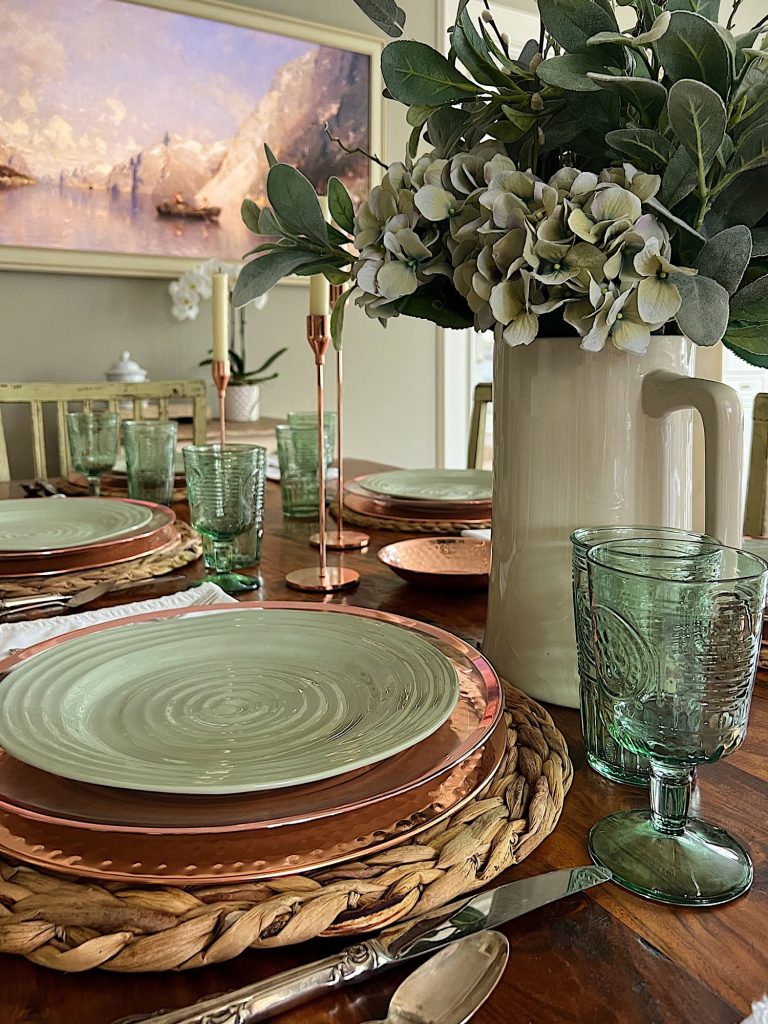 table set with hyacinth placemat, copper charger, green plate, green glasses, copper candlesticks, and green flowers in a vase. Frame TV in background.