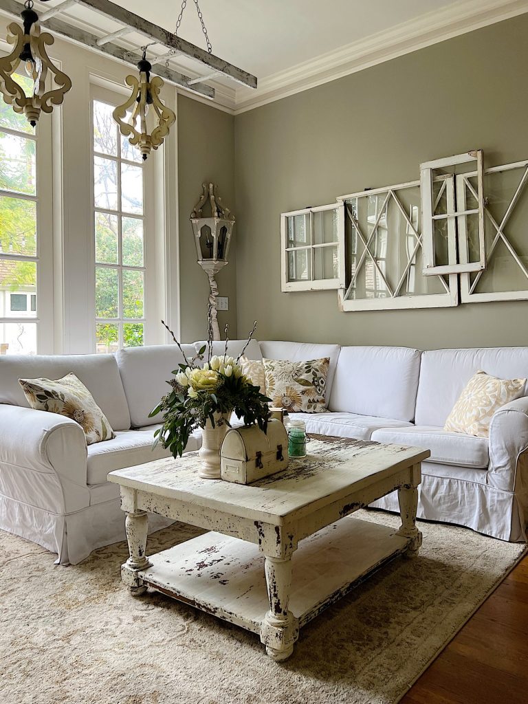 Family room with white section, hanging chandelier, vintage white coffee table, and decor items in sage green and taupe.