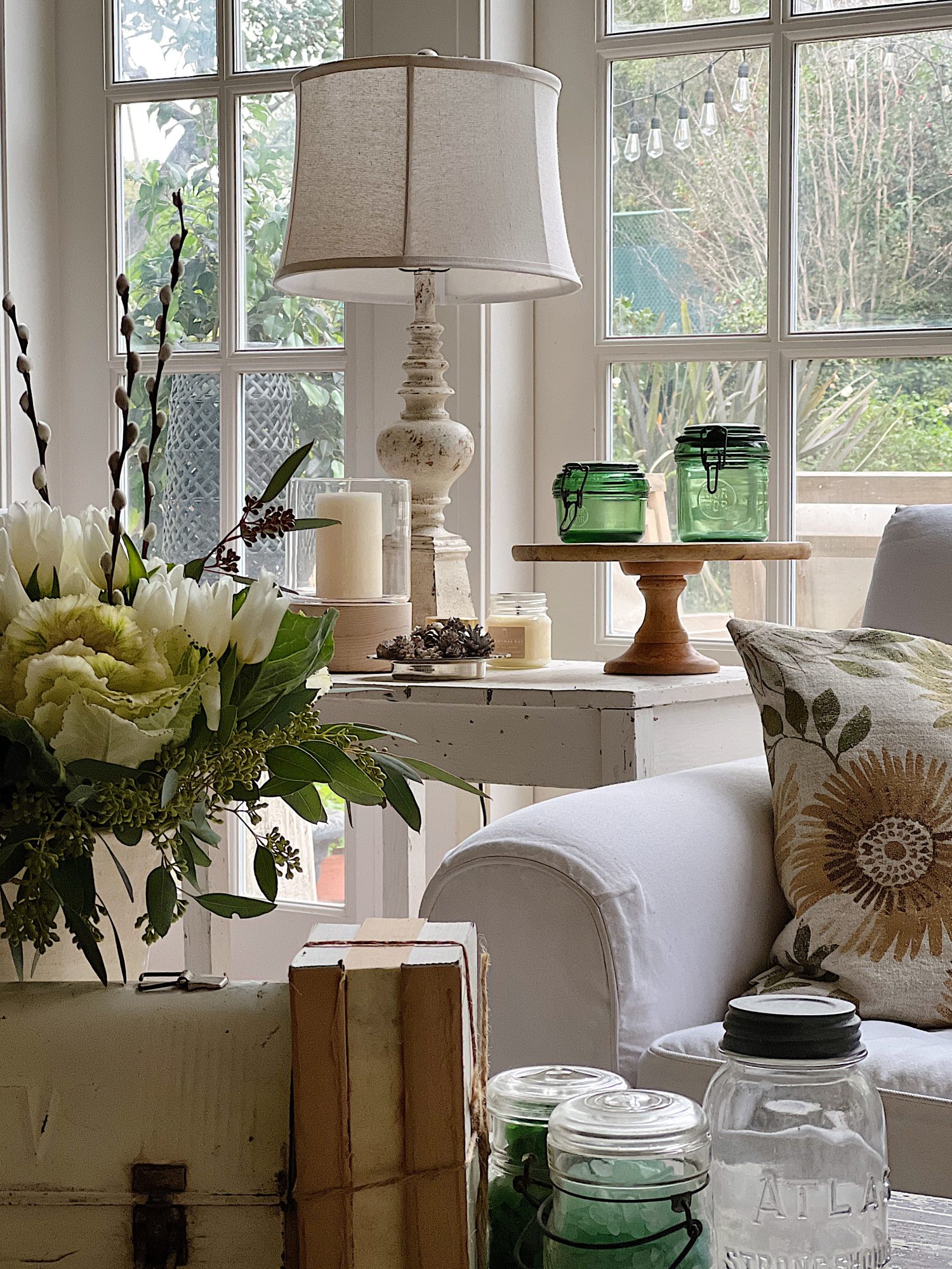 White couch with pillow and end table with lamp, green jars and candle. Coffee table with flowers, books, sea glass in jars and empty vintage glass far.