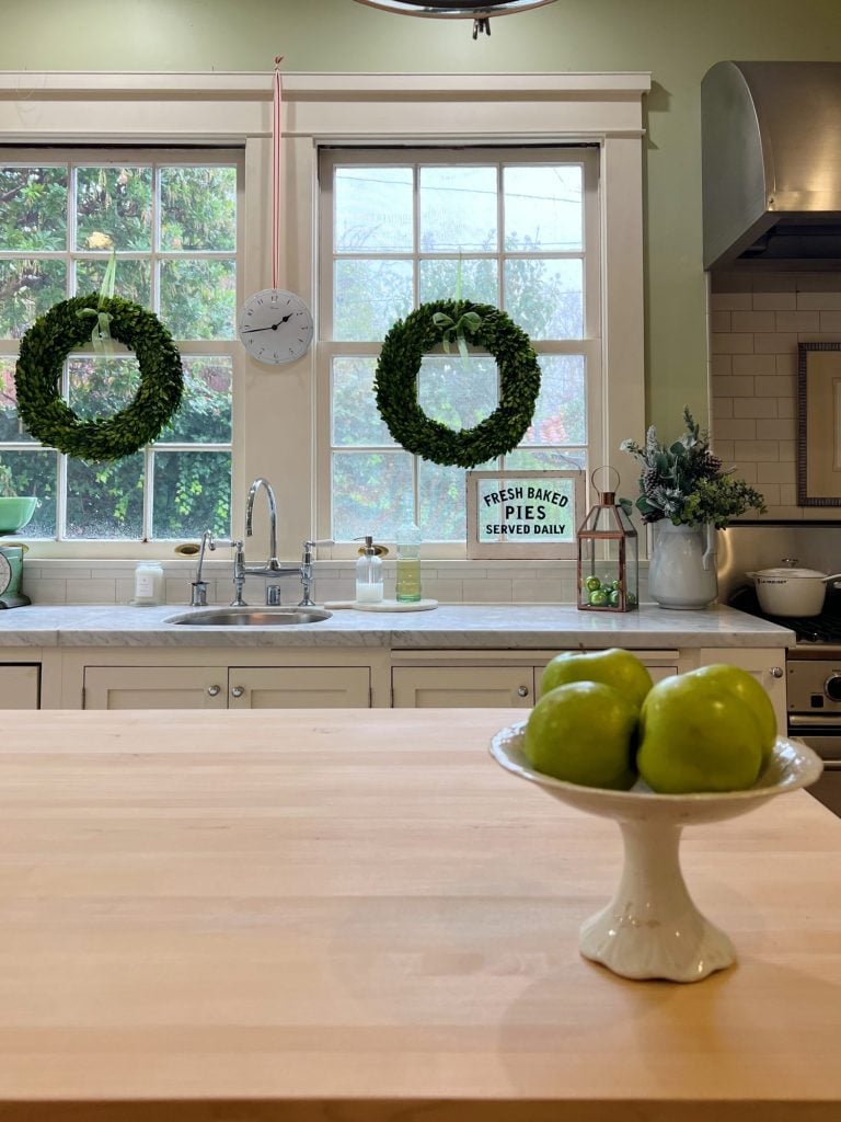 a white kitcehn with windows and an island with a bowl with apples, a cutting board and wreaths in the windows.