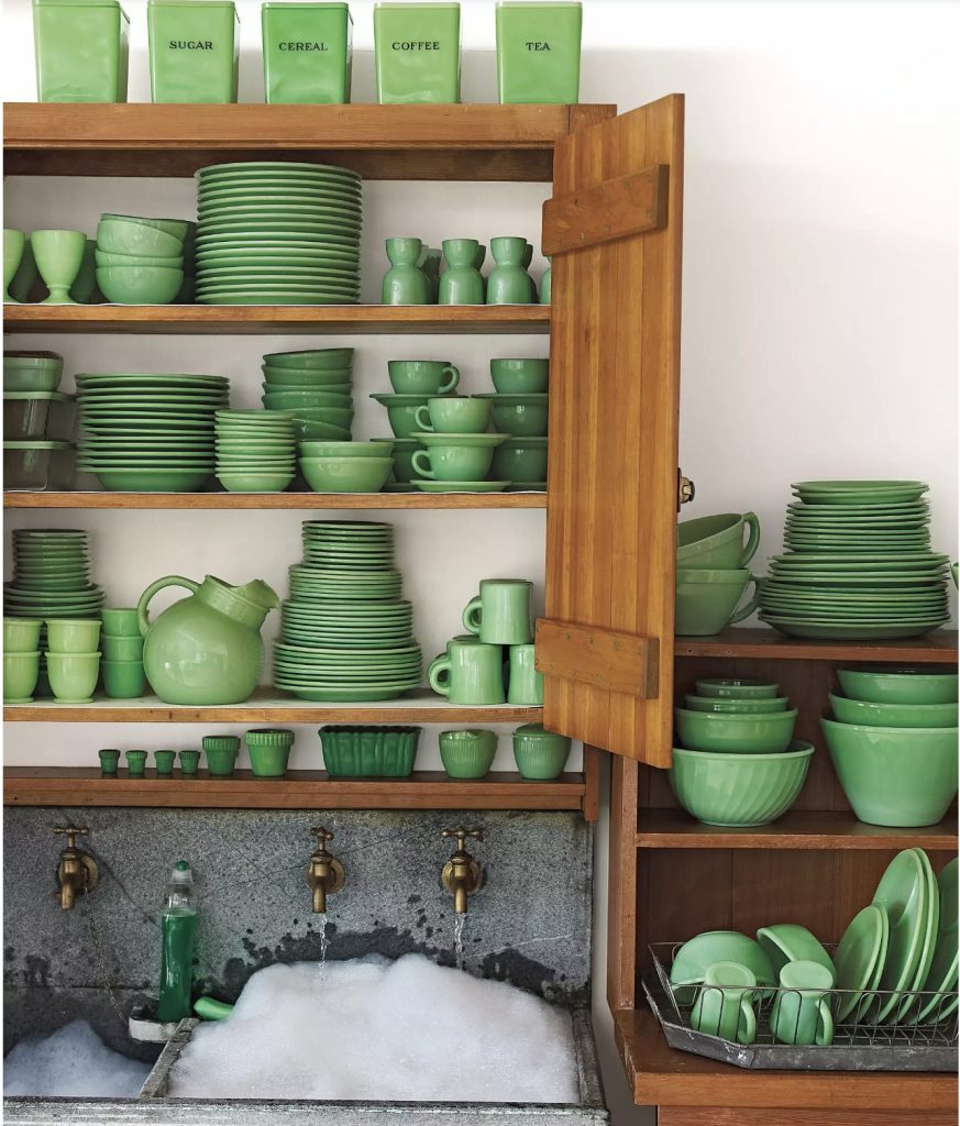 Wooden cupboard filled with jadeite cannisters, plates, bowls, pitchers and mugs surrounding a concrete sink filled with soap suds with three brass faucets