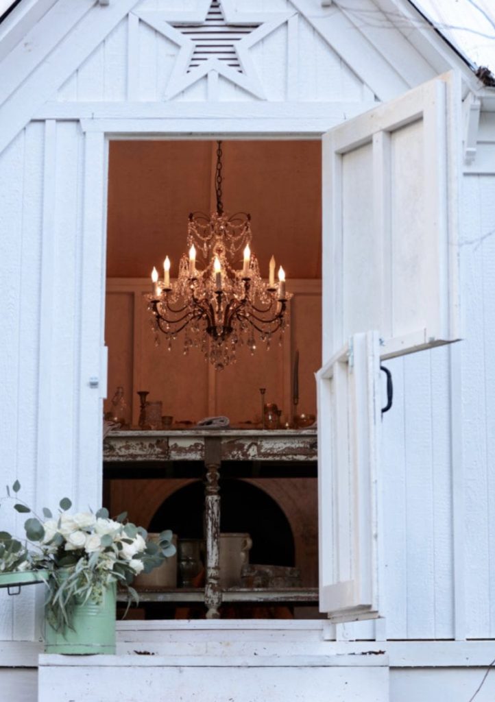 Open barn door with small green metal container filled with white flowers and eucalyptus on steps, leading to room with massive crystal chandelier and weathered table