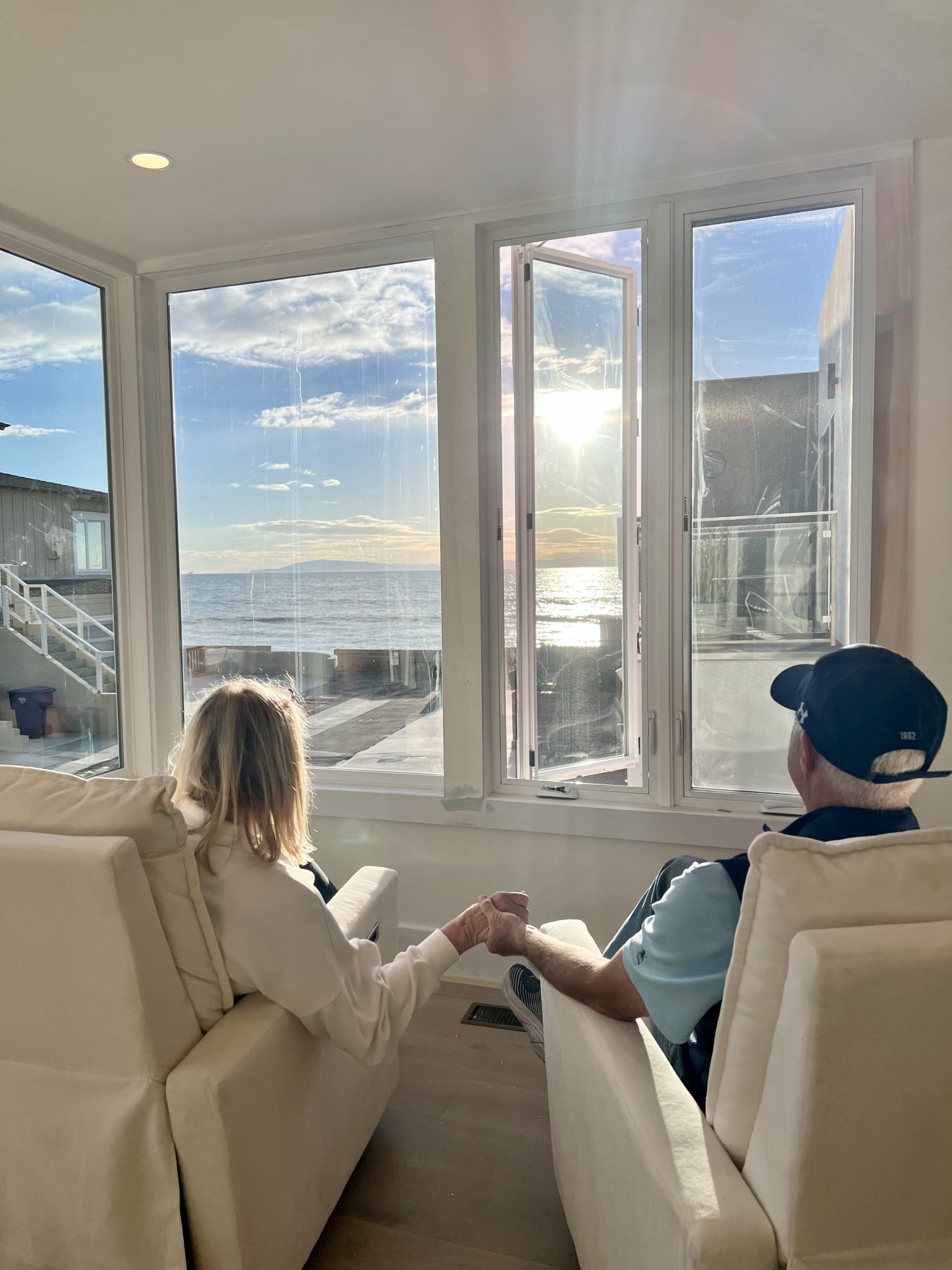 A couple sitting in white fabric covered chairs holding hands and gazing out the window at the ocean view