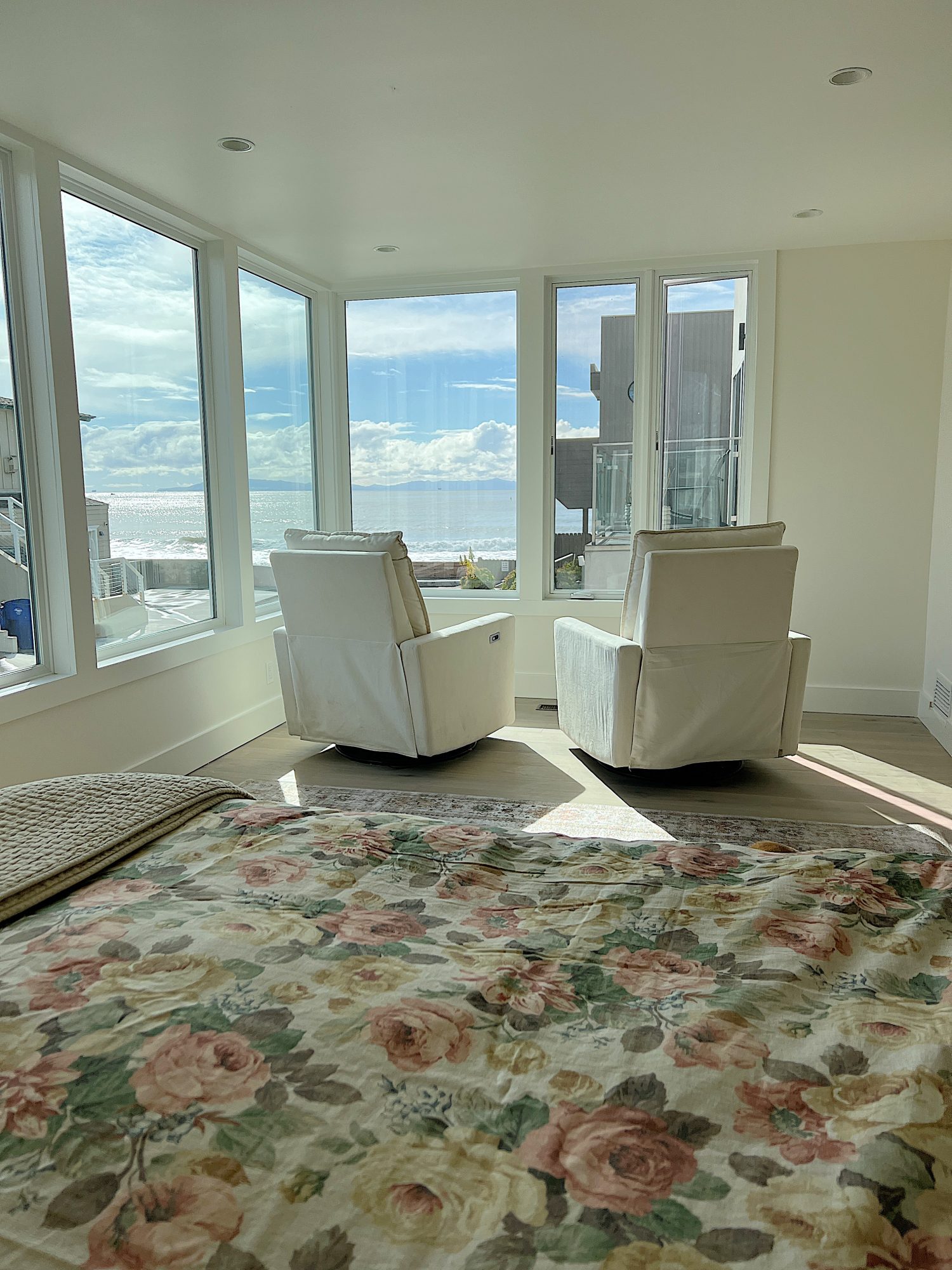 The primary suite with two a floral duvet and two chairs facing large windows with a view of the ocean.