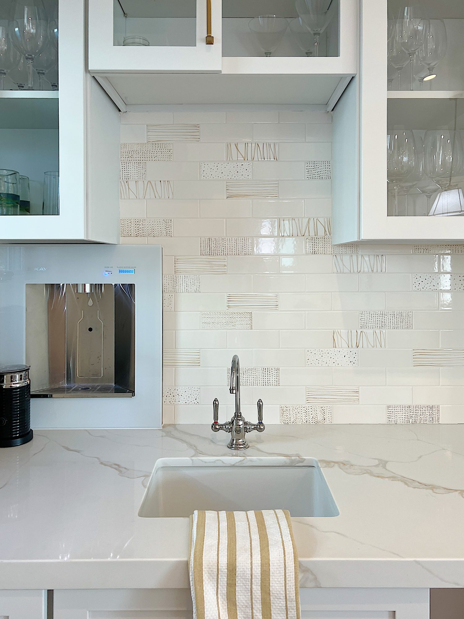 Beverage side of the kitceh with hand painted tiles, water dispenser, quartz countertops and white cabinets.