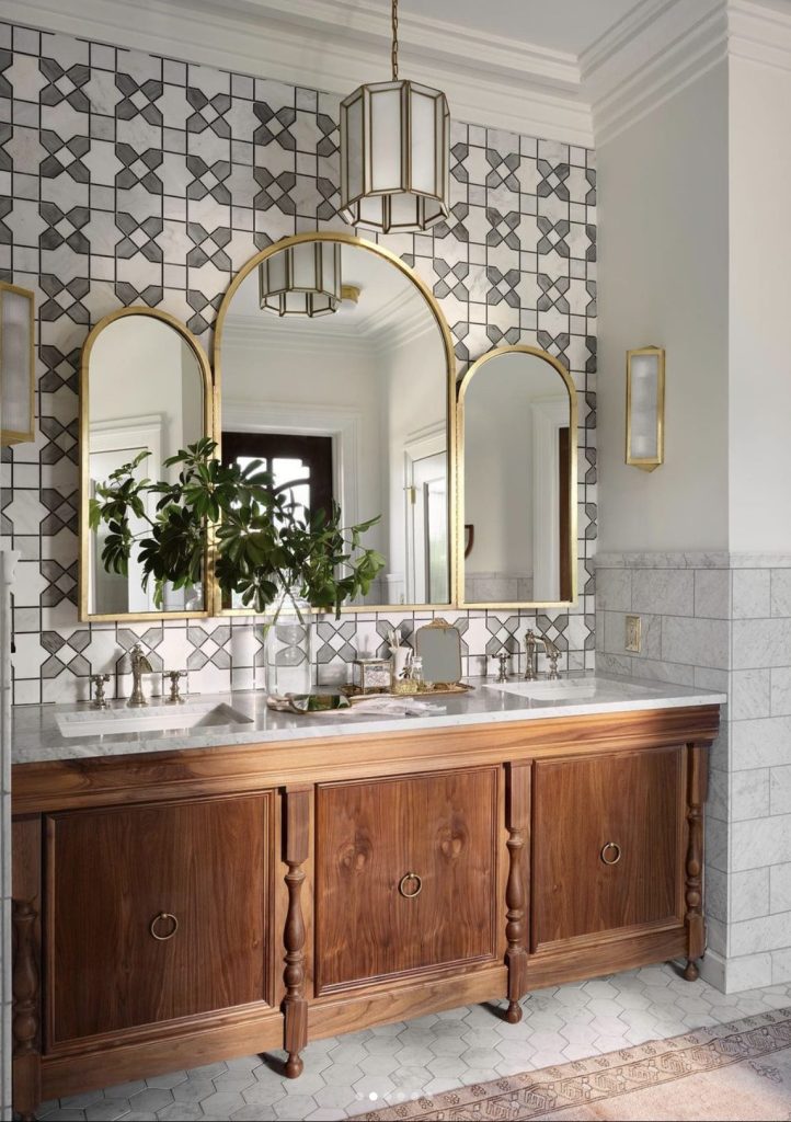 Bathroom at The Castle in Waco. Gray and white tile wall with three arched gold framed mirrors on top of walnut console holding two white sinks. A glass vase with greenery is in between the sinks