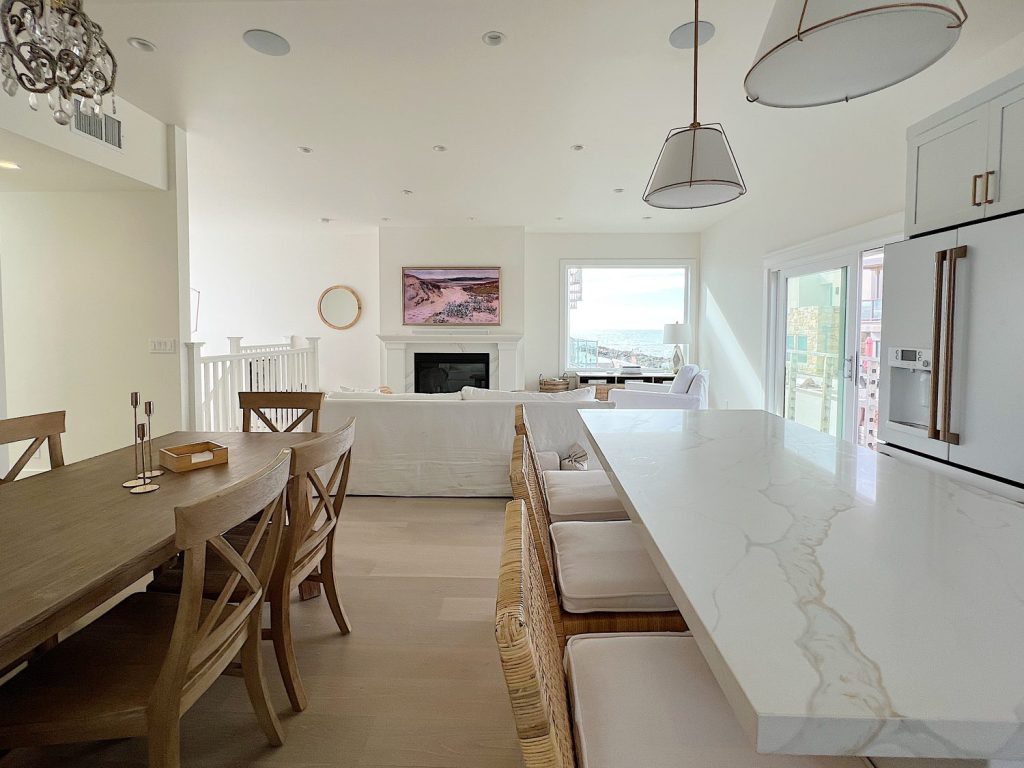 New beach house remodel living are and kitchen. White couch with pillow with white kitchen with white tile, quartz island, appliances, wood table and white cabinets.