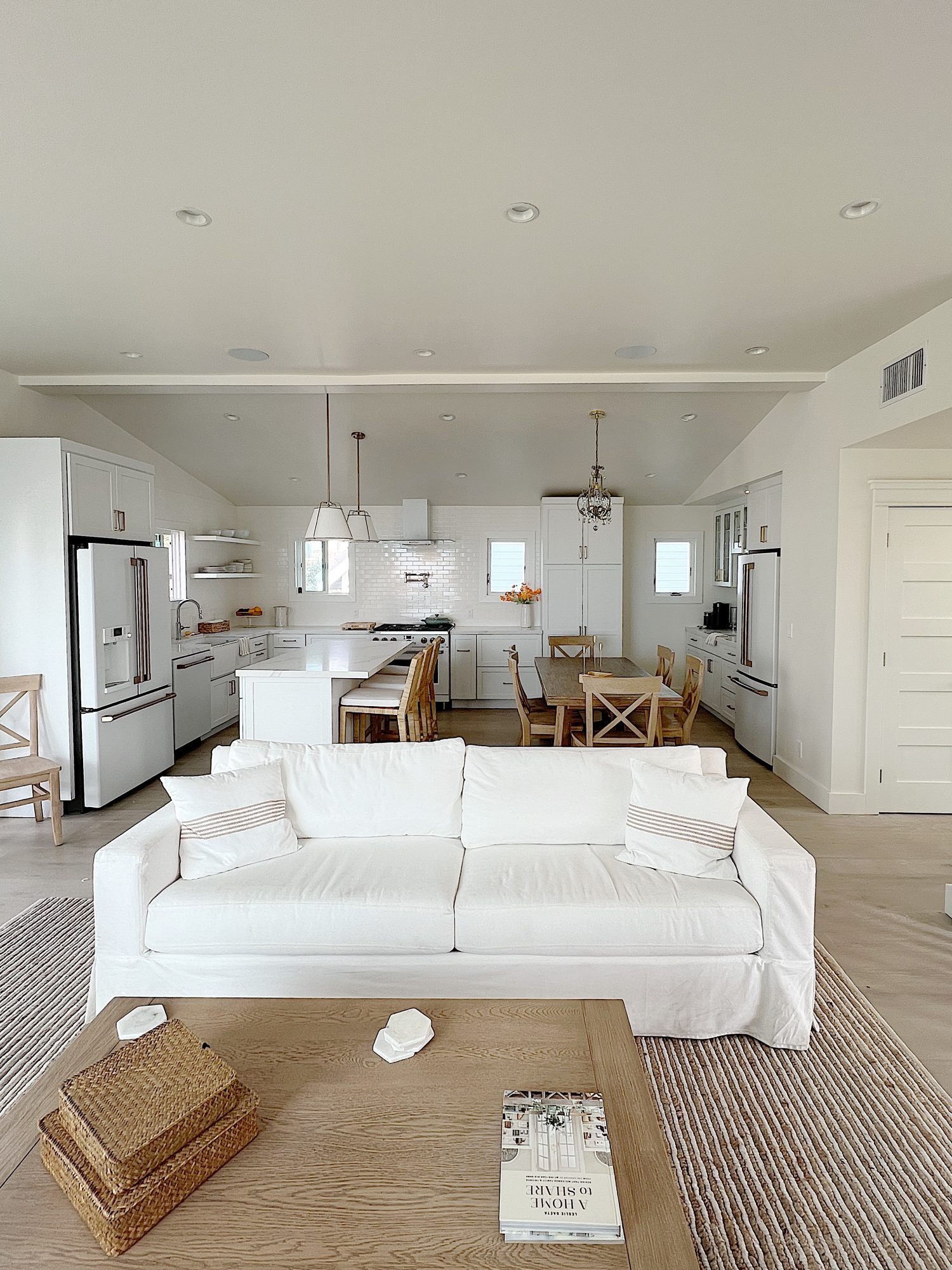 New beach house remodel living are and kitchen. White couch with pillow with white kitchen with white tile, quartz island, appliances, wood table and white cabinets.
