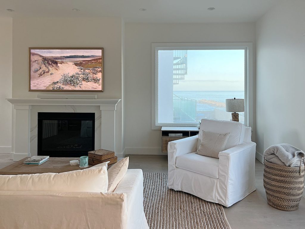Family living room at the beach house with white couch and chair, frame tv, and a window with a beautiful view.
