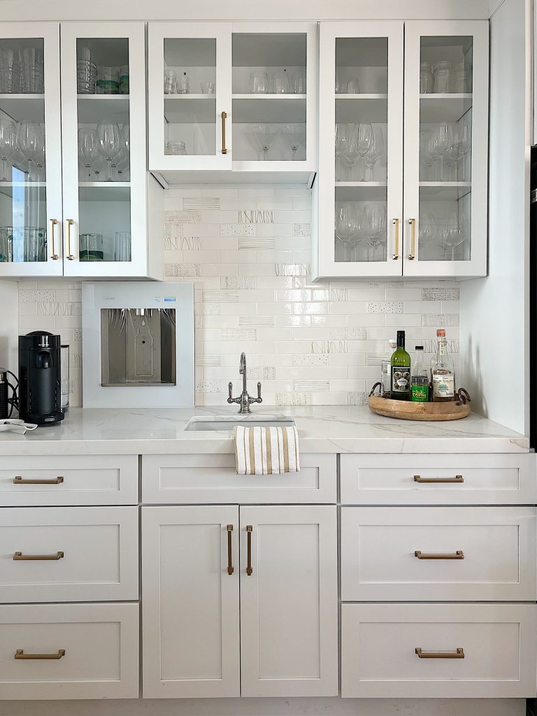 Kitchen beverage area with white cabinets, white quartz countertops, hand-painted tile, water dispenser, coffee maker, and towel.