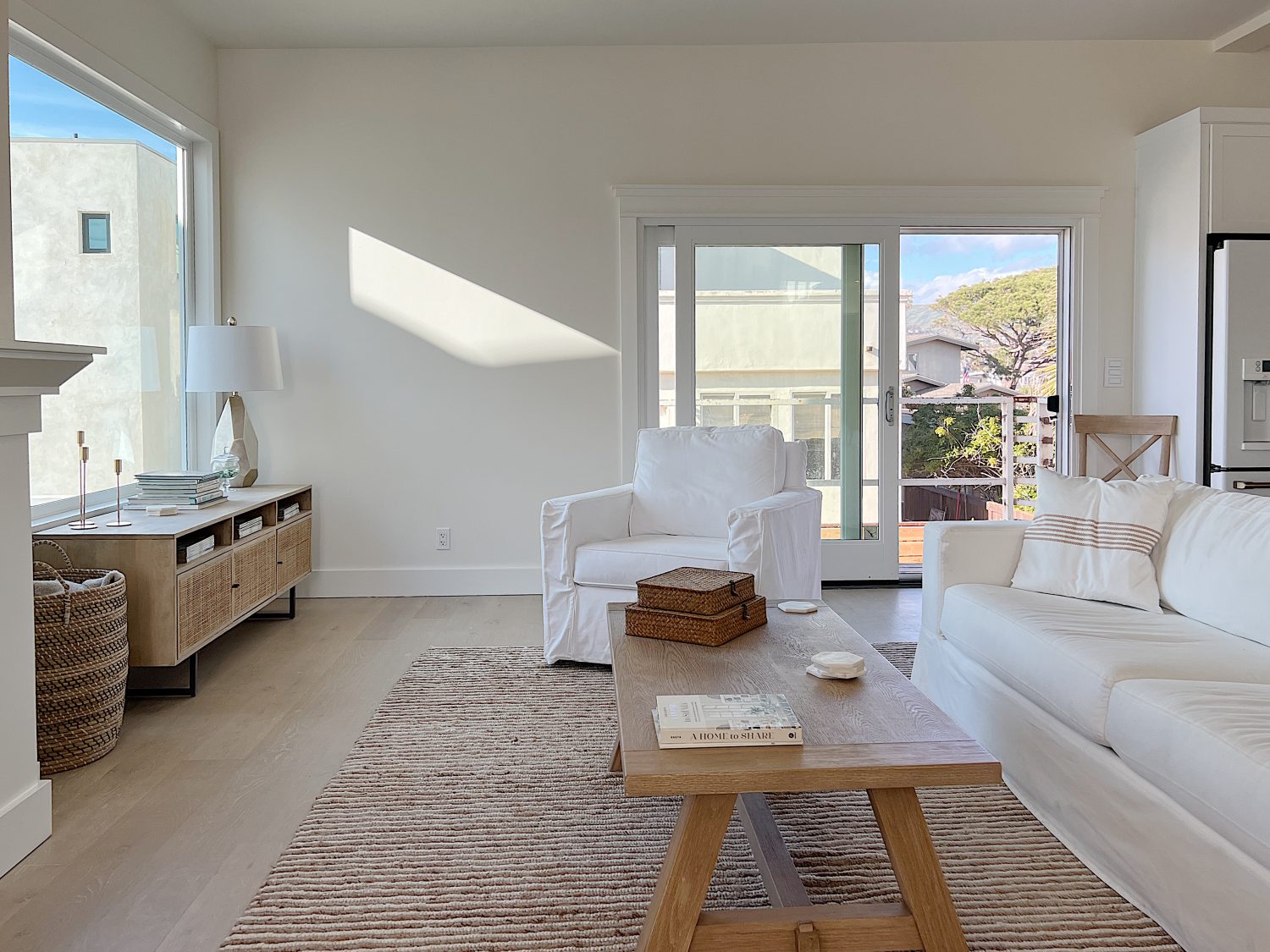 New beach house remodel living area with white couch with pillow, frame tv, rattan media console, basket, wool rug, light wood coffee table, white swivel chair and accessories.