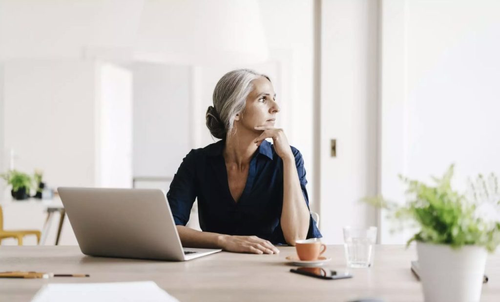 Woman working at a desk with open laptop, cup of coffee and glass of water. She is gazing out the window in concentration