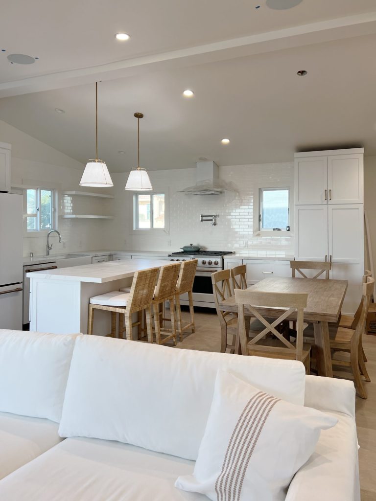 New beach house remodel living are and kitchen. White couch with pillow with white kitchen with white tile, quarts island, appliances, wood table and white cabinets.