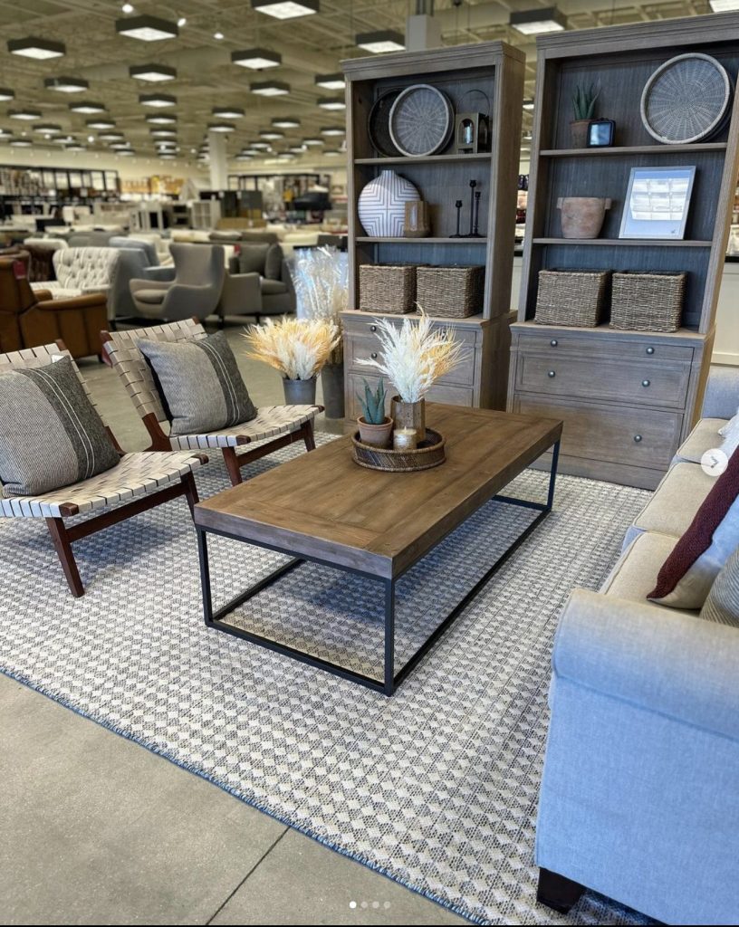 Styled bookcases, wood and iron coffee table, gray sofa and woven chairs on top of gray and white geometric rug. Background is rows and rows of outlet chairs and home items