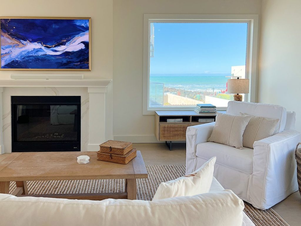living room of the beach house with a white couch and chair, a fireplace and two light wood tables. The fireplace has a frame TV above it with a beautiful photo.