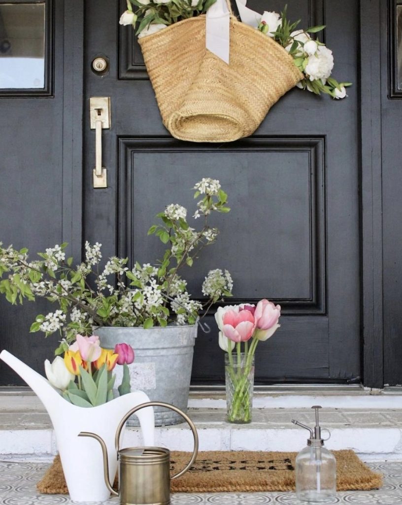 Close-up of black front door with a french market basket filled with white flowers is hanging from a fat white silk ribbon. On the doorstep is a galvanized bucket holding blooming dogwood branches. A criss-cut tall vase holds pink and white blooming tulips and a modern white watering cap holds yellow, pink and white tulips. A jute doormat and a small brass watering can and glass mister are also on the doorstep