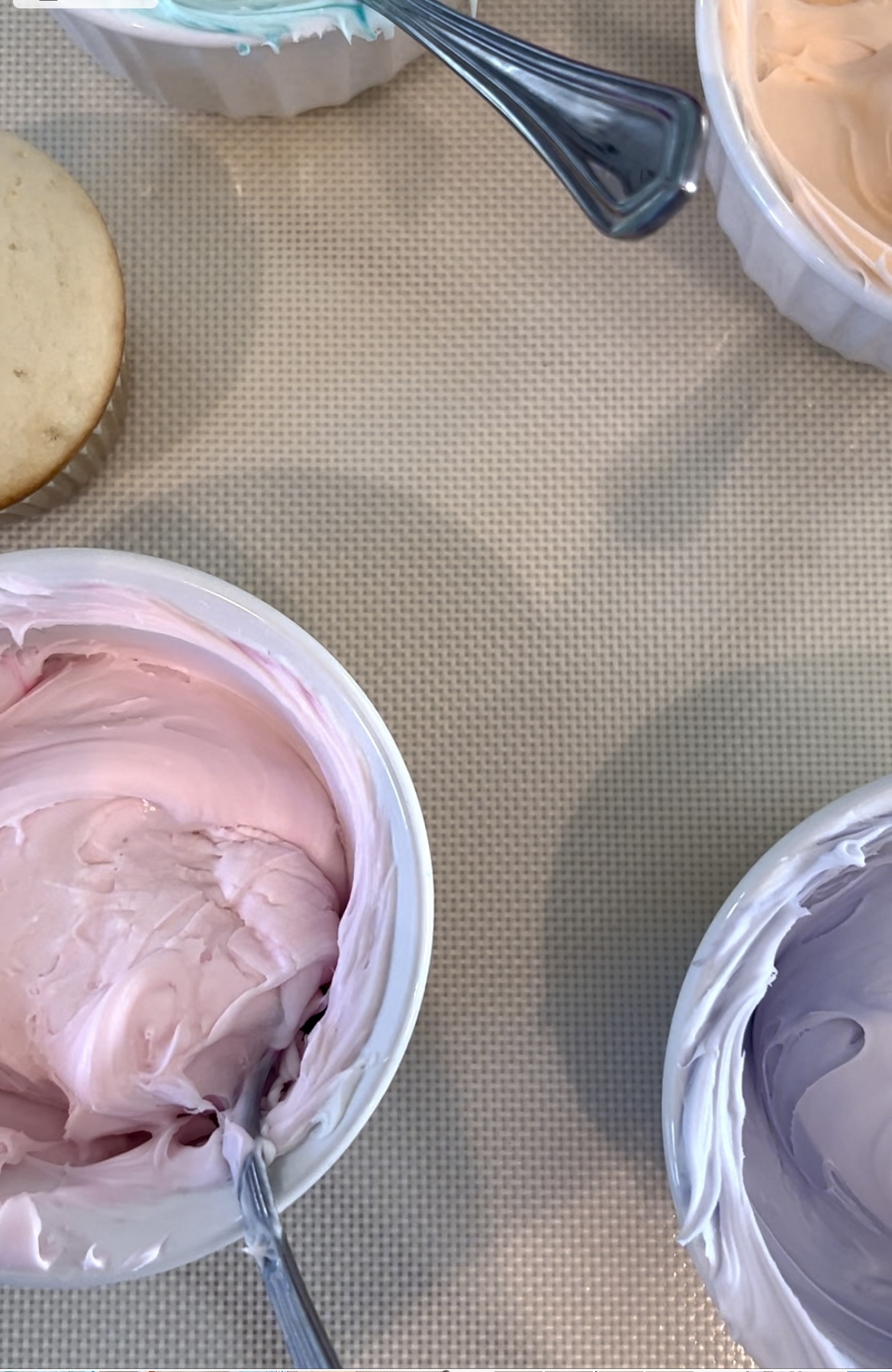 Silpat with four small ramekins filled with buttercream frosting in baby pink, lavender, mint green and orangecicle orange. Each of the bowls has a spoon in it also. There is an unfrosted cupcake to the left of the photo