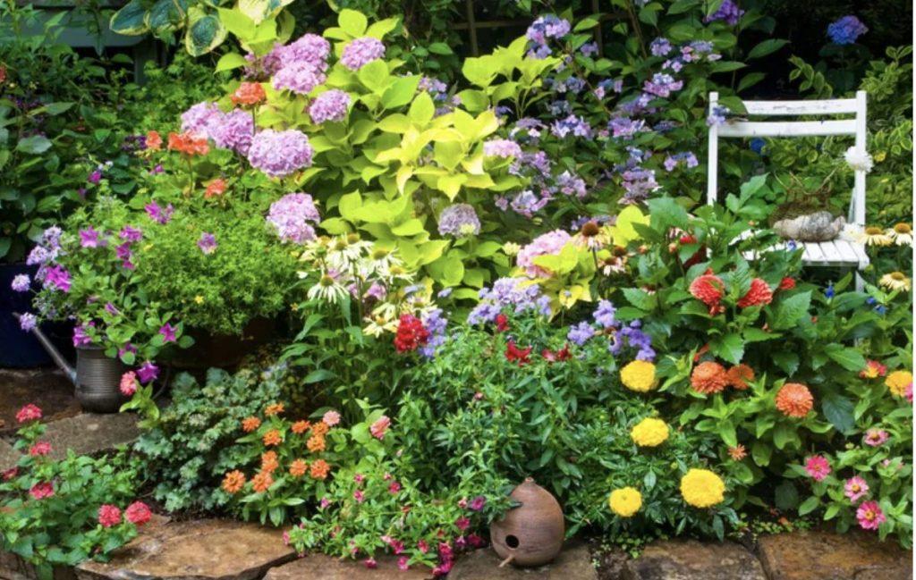 Gorgeous spring garden with colorful flowers and greenery. A shite wooden folding chair, a copper watering can and a clay bird house are tucked into the greenery.
