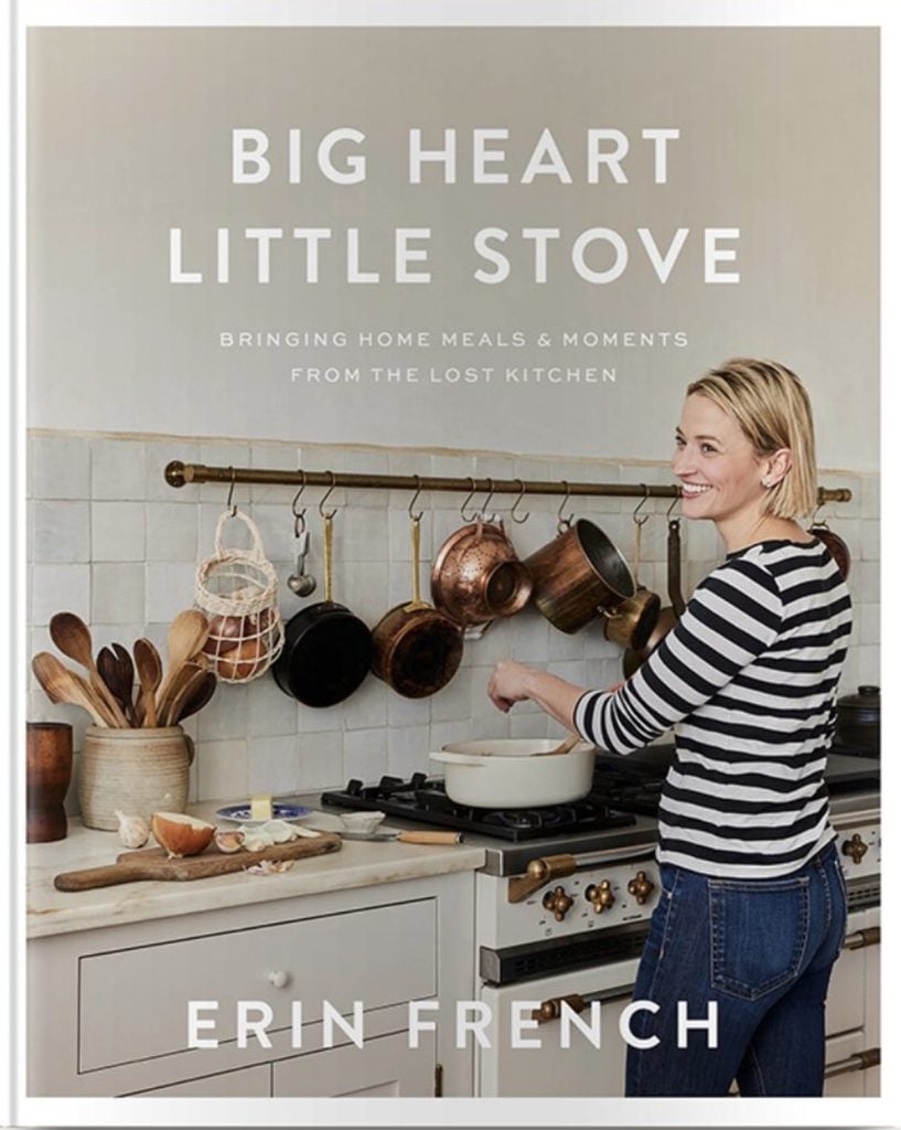 Cookcook cover, chef Erin French wearing blue and white striped shirt and jeans standing at a stove with pots hanging on the wall above the stove. Cookbook name is Big Heart Little Stove
