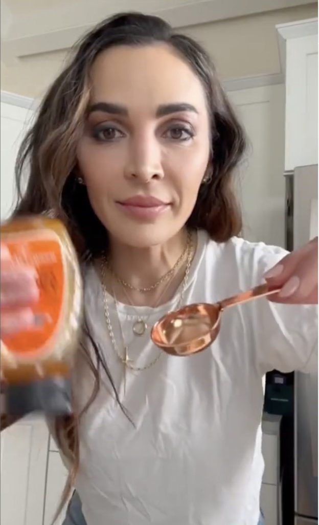 Beautiful woman with dark eyes and dark hair, wearing a white t-shirt and layered necklackes demonstrates how to measure honey in a copper measuring spoon