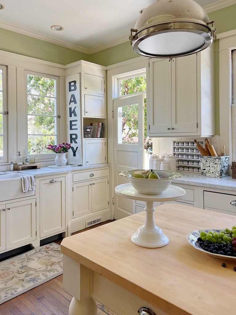 A traditional kitchen with white cabinets and light green walls and a butcher block kitchen island decorated with sage green and lavender items for spring.
