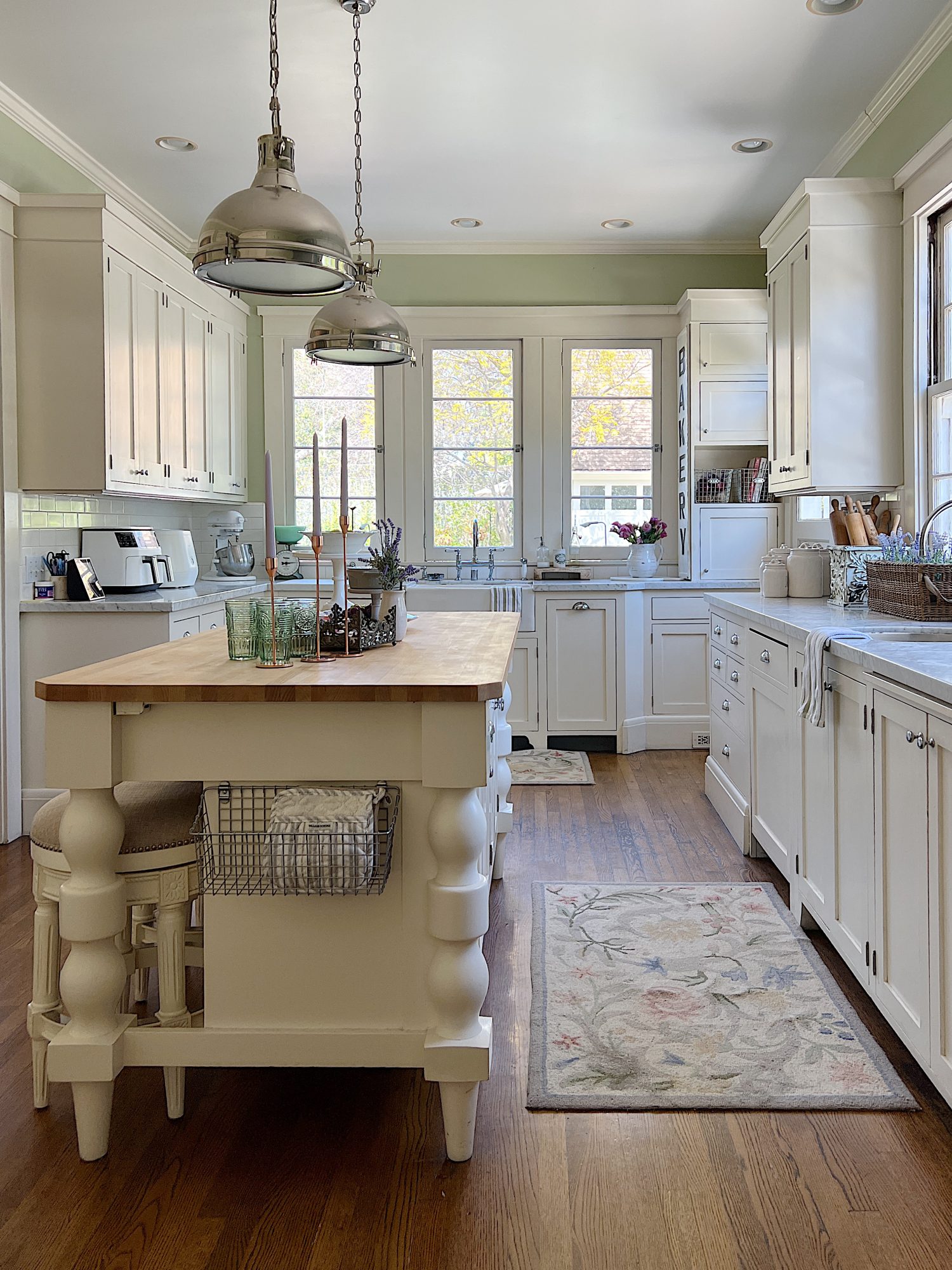 A traditional kitchen with white cabinets and light green walls and a butcher block kitchen island decorated with sage green and lavender items for spring.